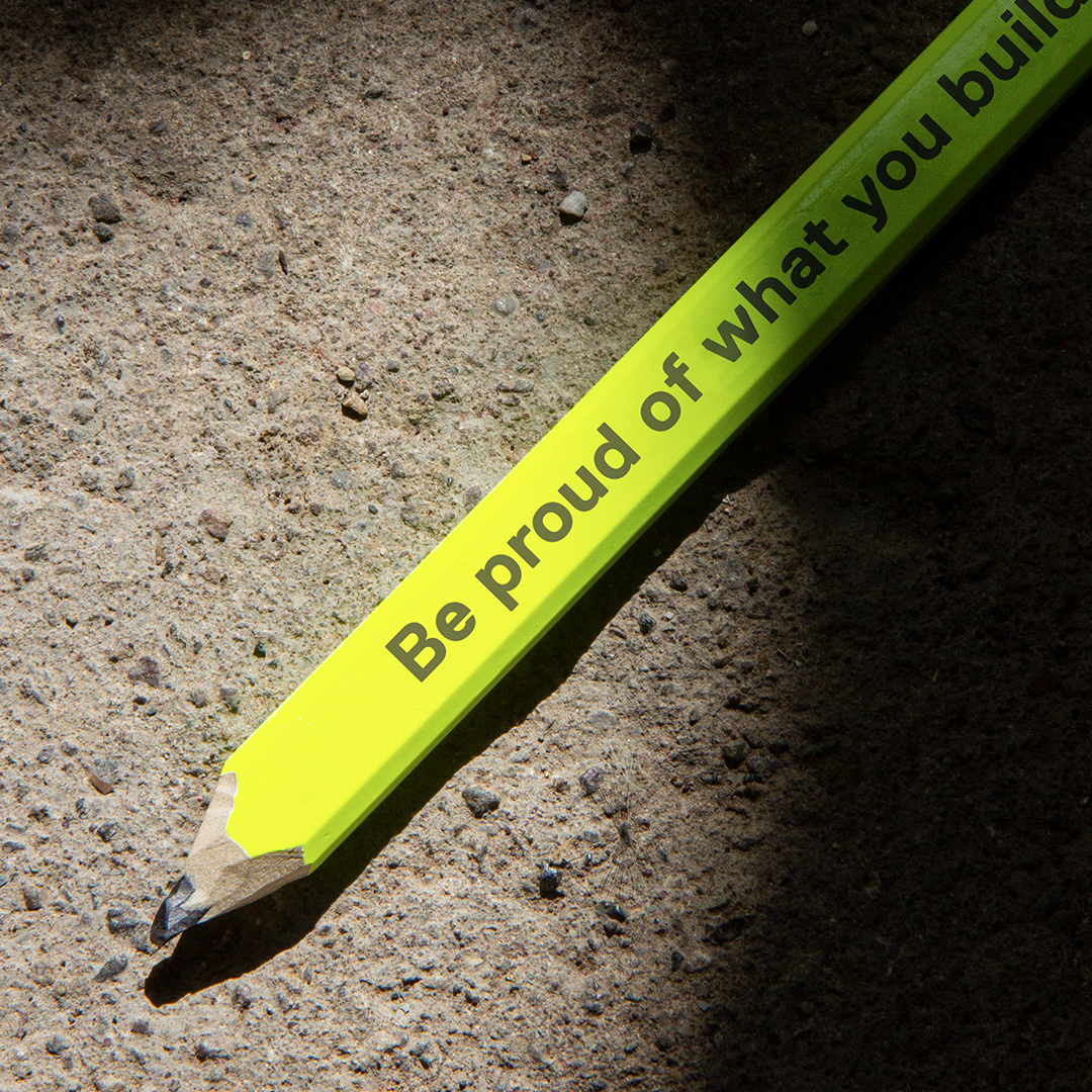 Neon-gelber Bleistift mit der Aufschrift "Be proud of what you build" auf Steinboden