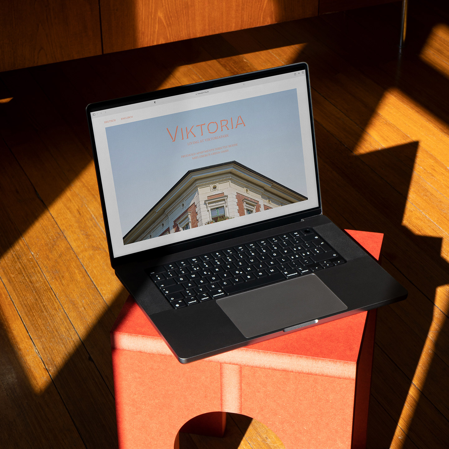MacBook steht aufgeklappt auf einem korall-farbenem Hocker, auf dem Screen ist die Startseite der "Viktoria" Website zu sehen mit dem Logo und einem großen Bild der Fassade