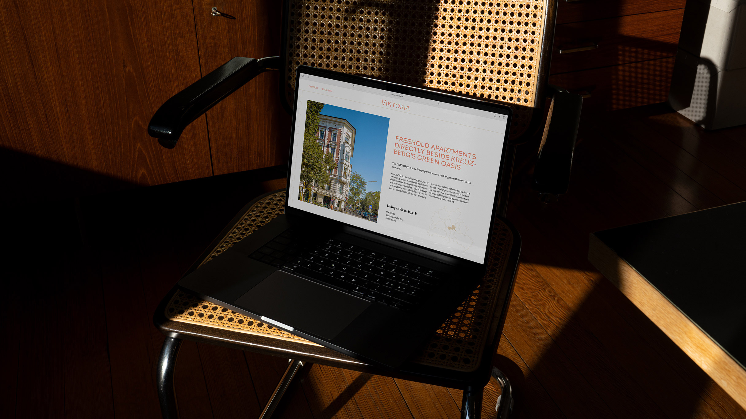 MacBook steht aufgeklappt auf einem korbgeflochtenem Stuhl, auf dem Screen ist ein Auschnitt der "Viktoria" Website mit einem Bild der Fassade und einem Text und einer illustrierten Karte über die Nachbarschaft und Lage des Hause